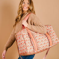 A smiling woman with long, wavy blonde hair in a tan sweater carries The Darling Effect’s Everbloom Dusty Rose On-the-Go Quilted Weekender Bag, featuring pink handles, over her shoulder against a beige background.
