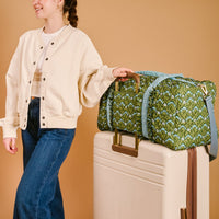 A person in a cream jacket and blue jeans stands next to a beige suitcase with The Darling Effect's Everbloom Forest Green On-the-Go Quilted Weekender Bag, featuring an adjustable shoulder strap, on top. They are smiling and holding the bag’s handle.
