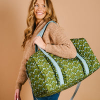 A smiling woman with long, wavy blonde hair carries The Darling Effect's Everbloom Forest Green On-the-Go Quilted Weekender Bag with light blue adjustable straps. She wears a tan knit sweater and blue jeans, against a plain beige background.