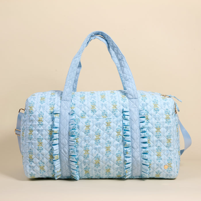 The Darling Effect Oh So Fancy Blue On-the-Go Quilted Weekender Bag features a light blue floral design, front ruffles, dual handles, a long adjustable strap, and a zippered top—spacious for travel—shown on a plain beige background.