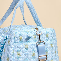 A close-up of The Darling Effect's Oh So Fancy Blue On-the-Go Quilted Weekender Bag, featuring a floral pattern, gold hardware, and matching blue straps, set against a beige background.