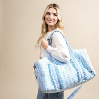 A smiling woman with long blonde hair carries The Darling Effect's Oh So Fancy Blue On-the-Go Quilted Weekender Bag, styled with ruffle details, over her shoulder. She wears a cream sweater and blue jeans against a beige background.