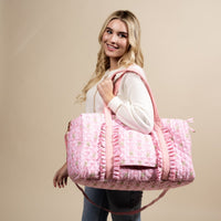 A smiling woman with long blonde hair carries The Darling Effect's Oh So Fancy Pink On-the-Go Quilted Weekender Bag on her shoulder while wearing a white sweater and jeans, standing against a beige background.