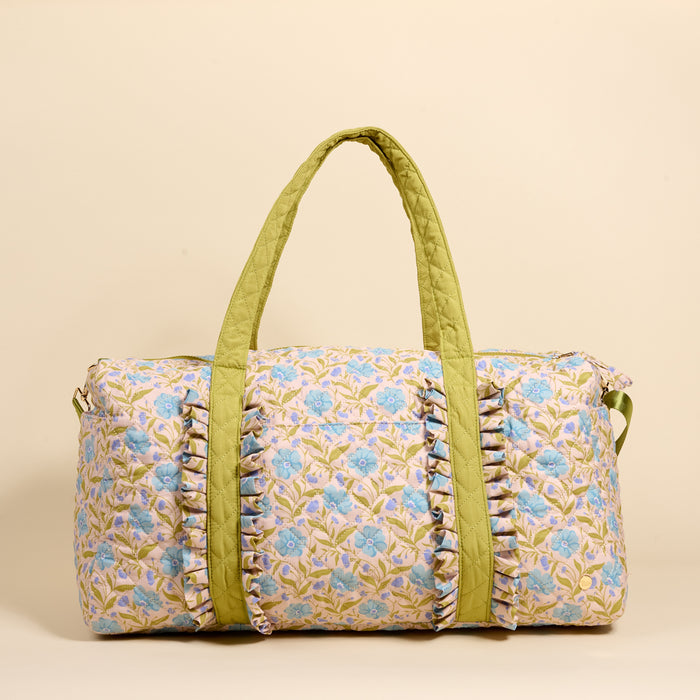 The Darling Effect’s Poppin' Petals Aqua On-the-Go Quilted Weekender Bag features a blue and green floral pattern, light olive green handles, and front ruffle details—ideal for travel. Shown against a beige background.