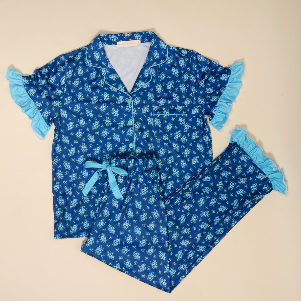 The Darling Effect Endless Daydream Navy Slumber Party Pajama Pants Set features a short-sleeve button-up top and matching pants with ultra-soft fabric, light blue ruffled trim, and a comfy waist tie on a beige background.