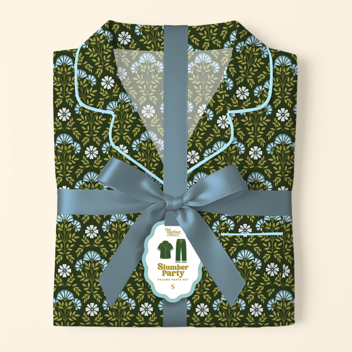 A folded Everbloom Forest Green Slumber Party Pajama Pants Set by The Darling Effect, in soft fabric with a floral print, is tied with a blue ribbon and bow. A tag shows the product name and a pajamas illustration.