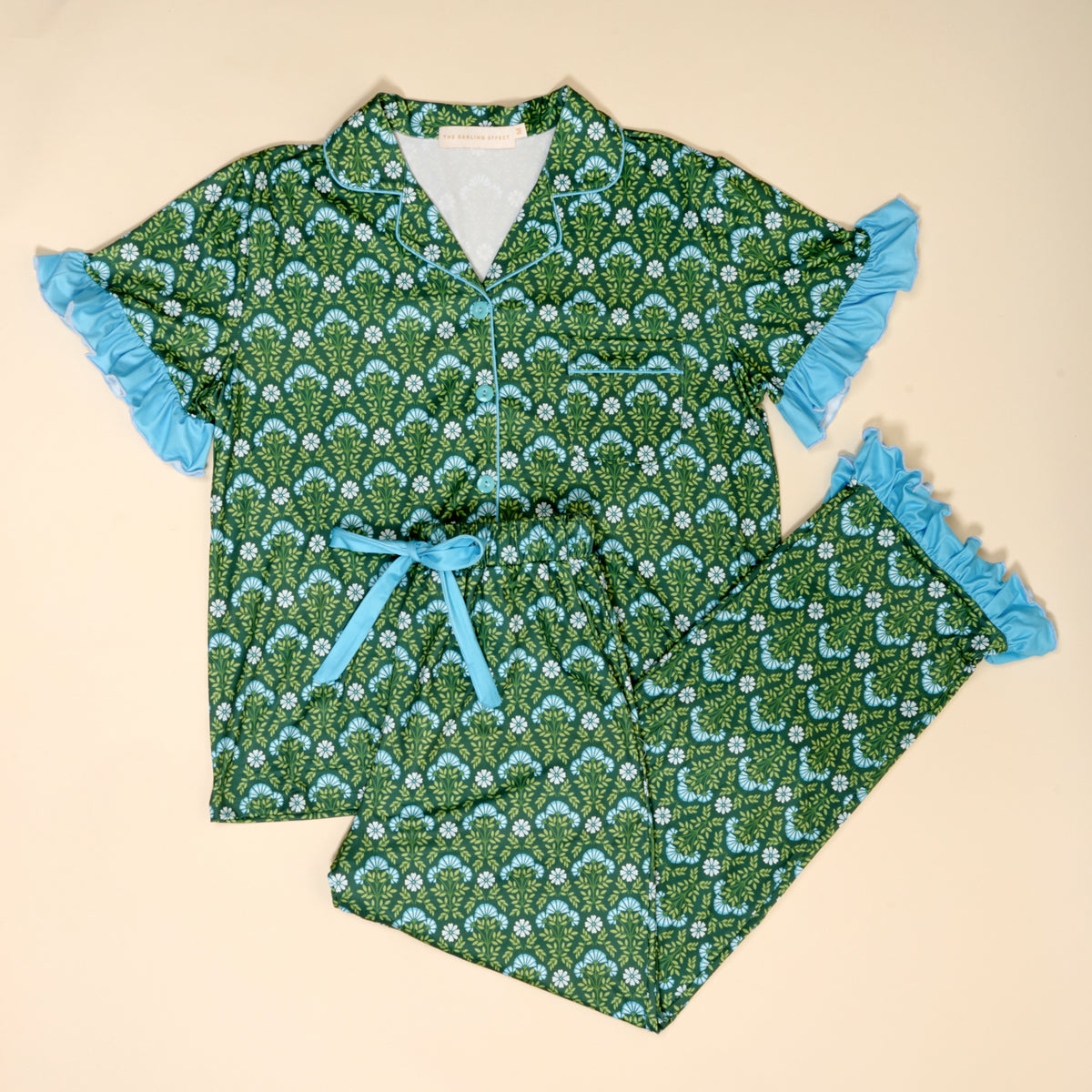 The Darling Effect Everbloom Forest Green Slumber Party Pajama Pants Set, featuring a leafy pattern, blue ruffled trim, chest pocket, and waist tie in a relaxed fit, is laid flat on a cream background.