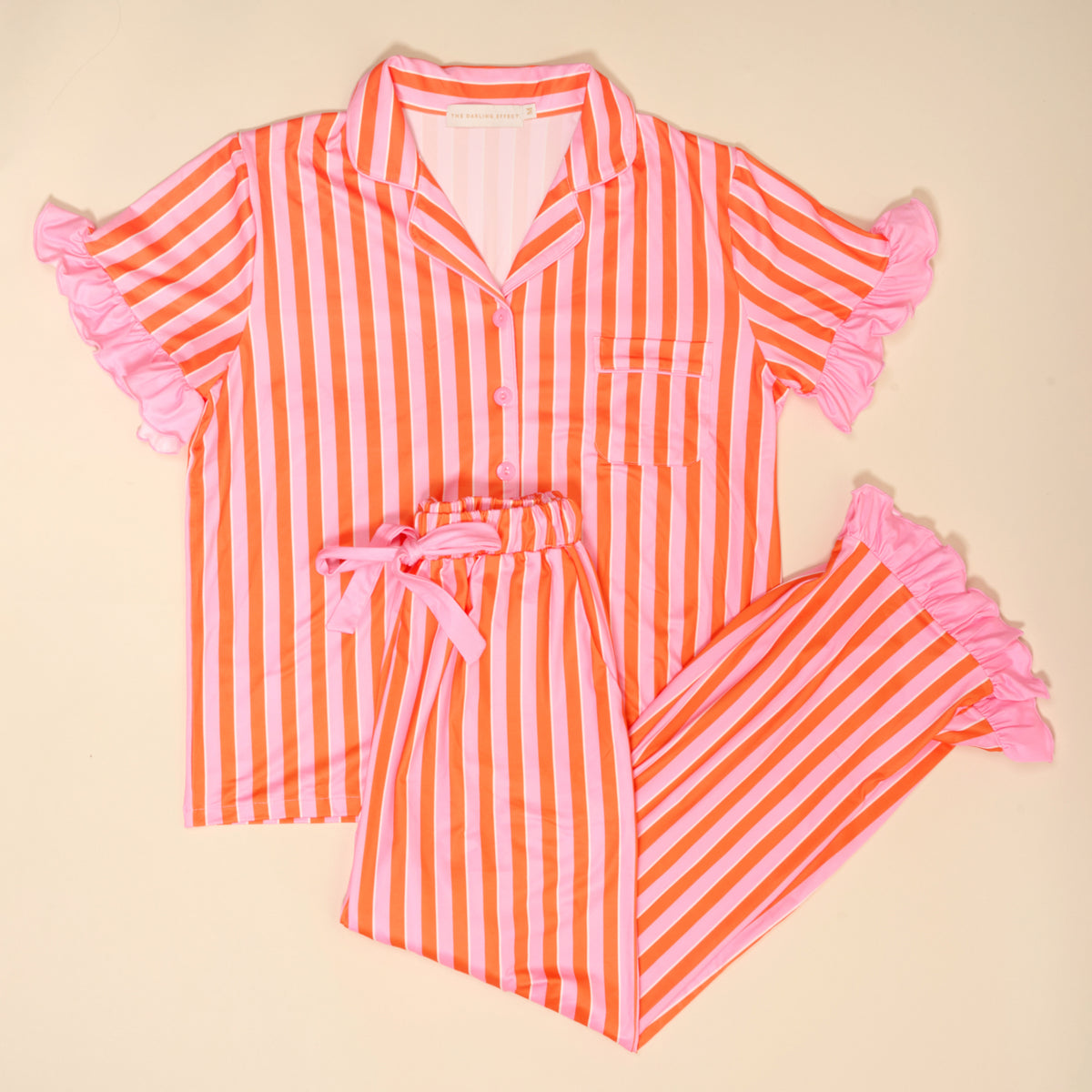 The Darling Effect Sicily Stripes Pink Slumber Party Pajama Pants Set features short sleeves, orange and pink vertical stripes, pink ruffle trim on sleeves and pant cuffs, crafted from ultra-soft fabric and shown on a light background.