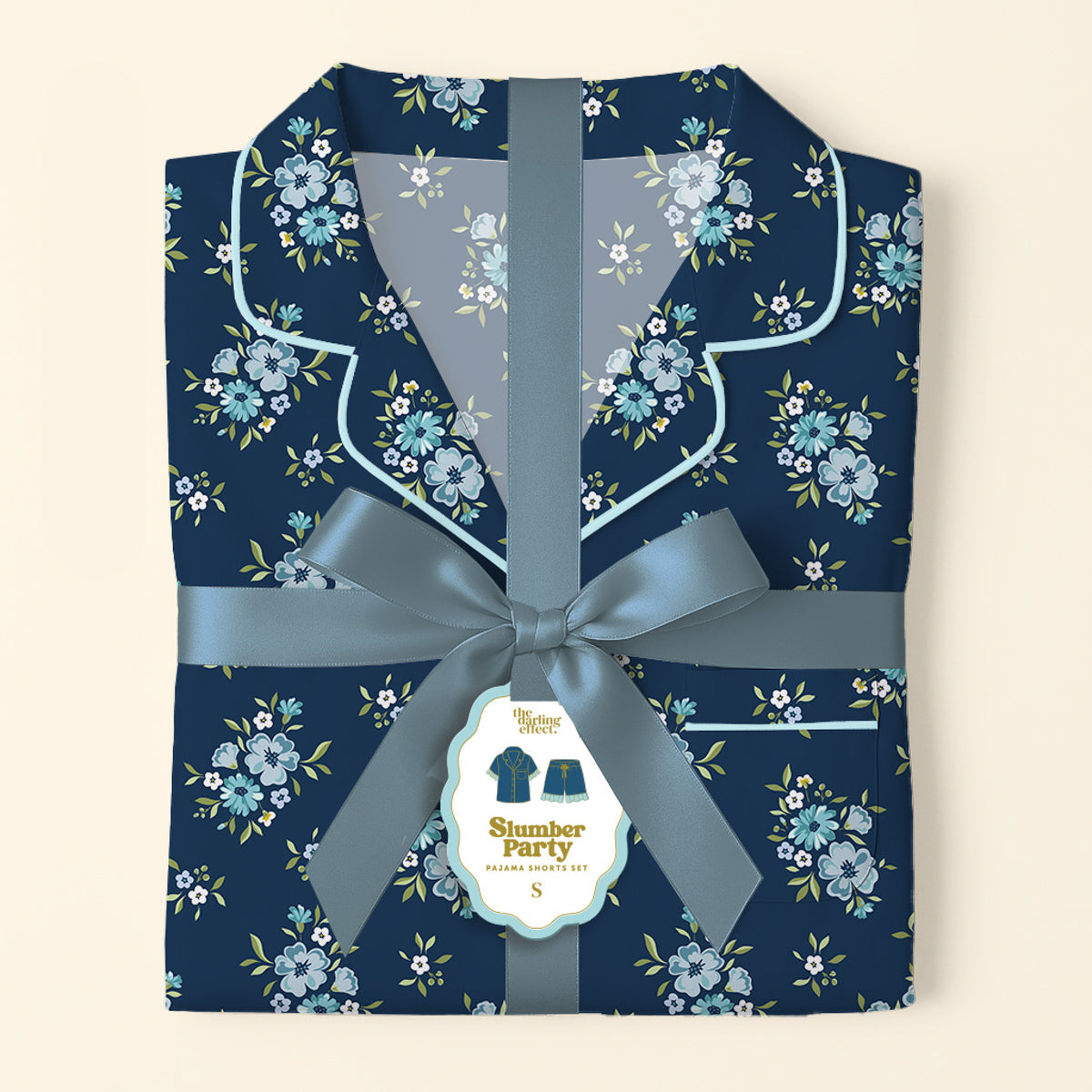 The Darling Effect’s Endless Daydream Navy Slumber Party Pajama Shorts Set features folded, ultra-soft navy pajamas with a light blue floral pattern, tied with a satin ribbon and bow. Gift-ready, it includes a tag with an illustration of the set.