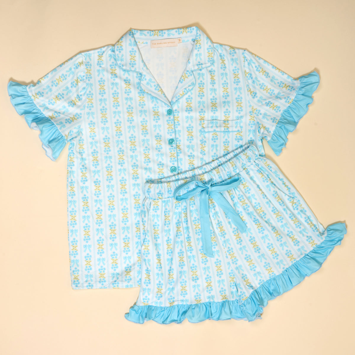 The Darling Effect's Oh So Fancy Blue Slumber Party Pajama Shorts Set features a short-sleeve button-up top and shorts with a light blue floral pattern and ruffled edges, made from ultra-soft fabric. Shown laid flat on a beige background.