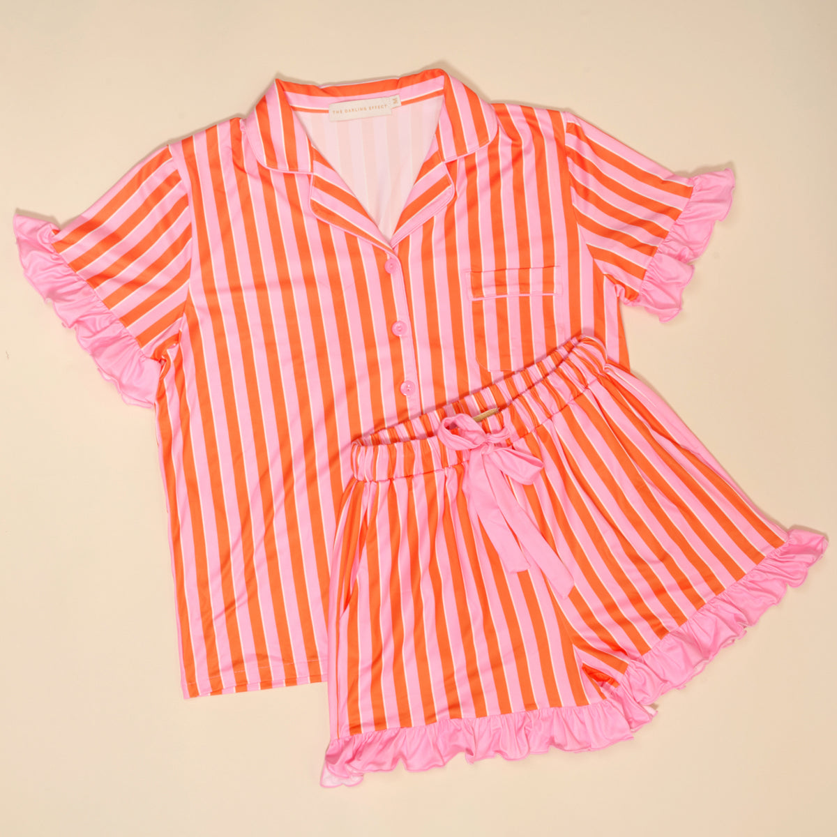 The Darling Effect’s Sicily Stripes Pink Slumber Party Pajama Shorts Set features pink and orange vertical stripes, ruffled hems, a front pocket, matching shorts with a pink ribbon tie—shown laid flat on a beige background.