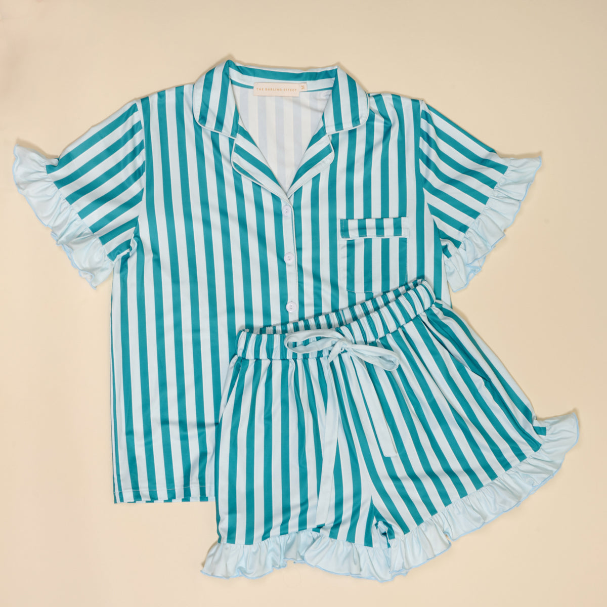 The Darling Effect’s Sicily Stripes Teal Slumber Party Pajama Shorts Set features teal and white stripes on ultra-soft fabric, with a short-sleeved ruffled button-up top and high-waisted shorts with a tie belt and ruffled hems, laid flat on beige.