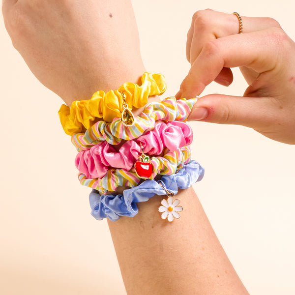 A forearm wears The Darling Effect’s Color Me Happy Scrunchie Charm Set—yellow, multicolored, pink, and blue scrunchies with teardrop, strawberry, heart, and daisy charms—as a hand touches these stylish hair accessories.