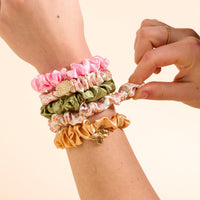 The Darling Effect's Endless Daydream Cream Scrunchie Charm Set features a satin scrunchie with hair charms, worn on the wrist and another being pulled gently by a finger.