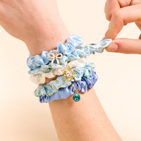 A hand displays the Oh So Fancy Blue Scrunchie Charm Set by The Darling Effect—blue and white floral scrunchies adorned with gold charms, including a bow, crystal heart, and teal teardrop—against a beige background.