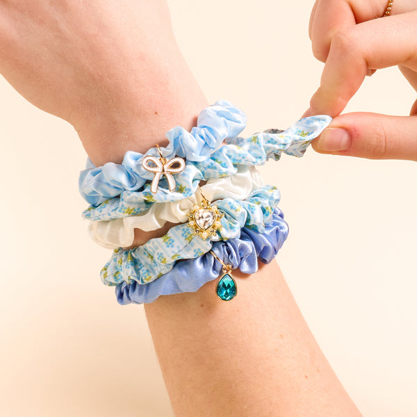 A hand displays the Oh So Fancy Blue Scrunchie Charm Set by The Darling Effect—blue and white floral scrunchies adorned with gold charms, including a bow, crystal heart, and teal teardrop—against a beige background.
