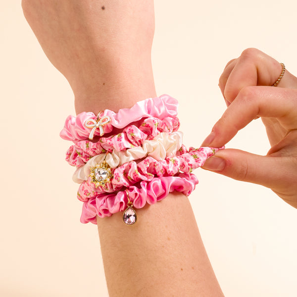 A hand is adorned with several scrunchies from The Darling Effect’s Oh So Fancy Pink Scrunchie Charm Set, featuring pink and white floral designs and small, chic hair charms. Another hand adjusts one of the stylish accessories against a plain light beige background.