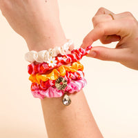 A hand showcases The Darling Effect's Sicily Stripes Pink Scrunchie Charm Set—stylish pink scrunchies with decorative charms—against a light beige background.
