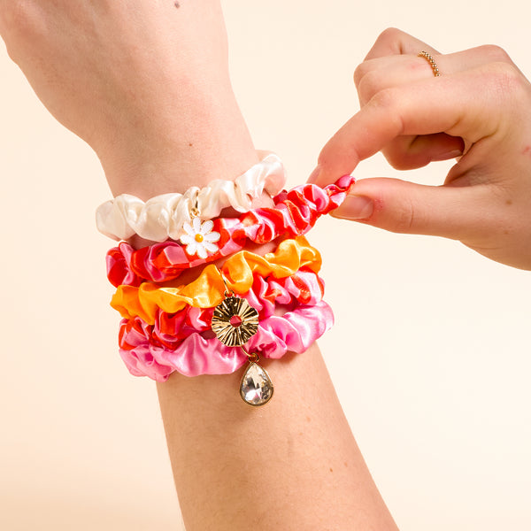 A hand showcases The Darling Effect's Sicily Stripes Pink Scrunchie Charm Set—stylish pink scrunchies with decorative charms—against a light beige background.