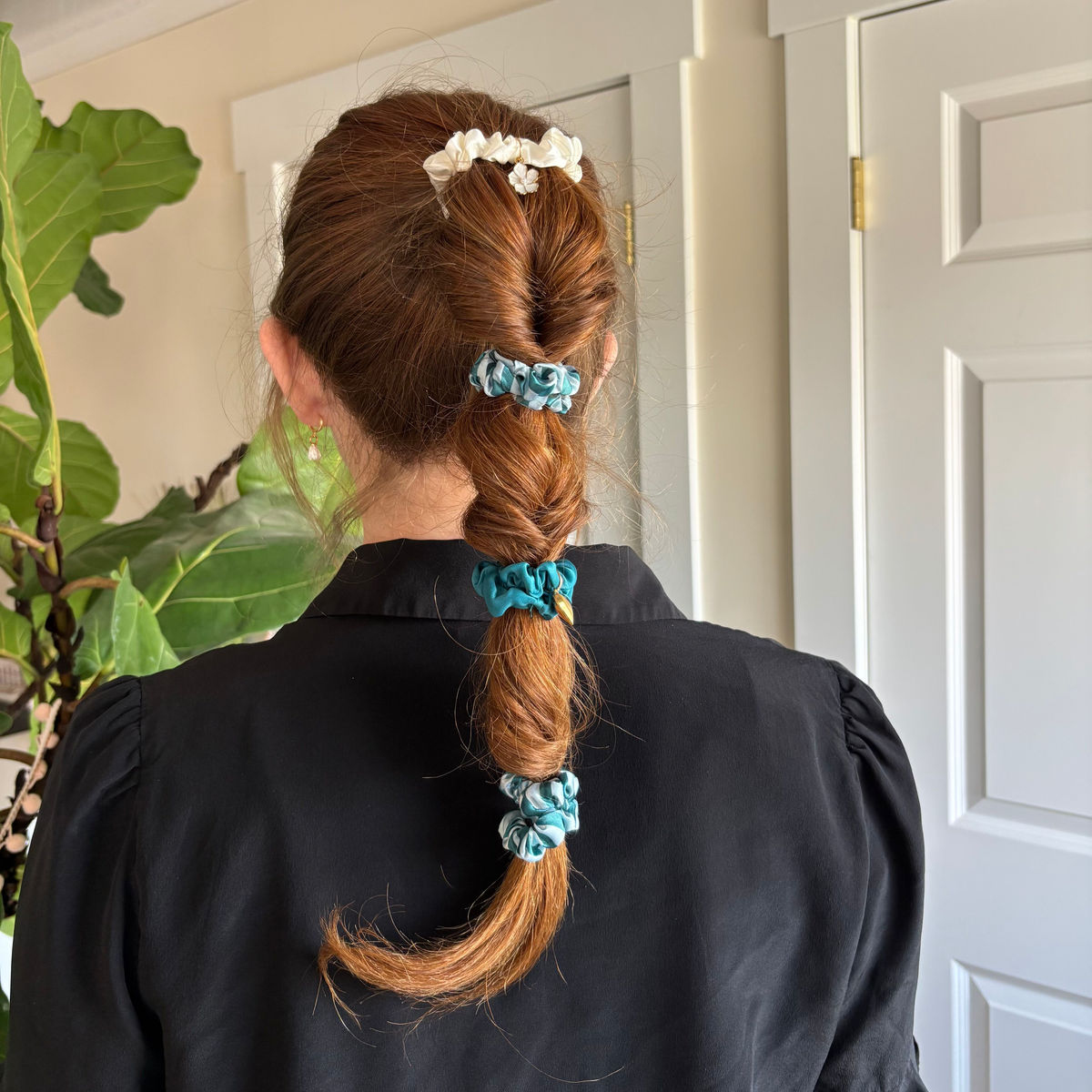 A person with reddish-brown hair in a segmented ponytail, styled using The Darling Effect’s Sicily Stripes Teal Scrunchie Charm Set, stands facing away in a black blouse; green leafy plants and white paneled doors are visible behind them.