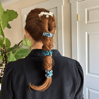 A person with reddish-brown hair in a segmented ponytail, styled using The Darling Effect’s Sicily Stripes Teal Scrunchie Charm Set, stands facing away in a black blouse; green leafy plants and white paneled doors are visible behind them.