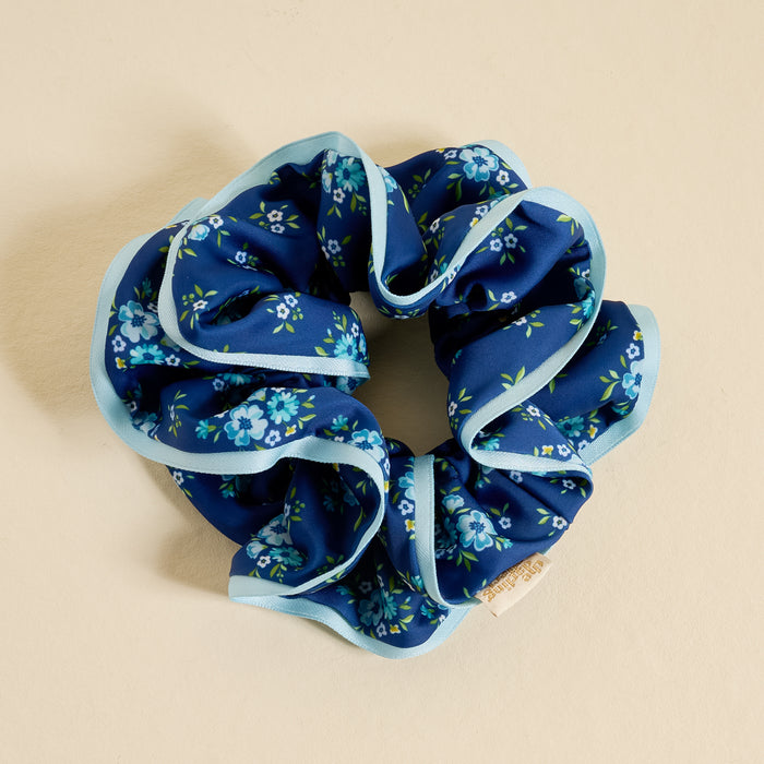The Darling Effect Endless Daydream Navy Ruffle Up Scrunchie features oversized blue satin with light blue trim and a floral pattern of small blue flowers and green leaves, styled in a circle on a beige background.