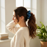 A woman with long brown hair tied in The Darling Effect’s Endless Daydream Navy Ruffle Up Scrunchie stands in a sunlit room, wearing a cream sweater, surrounded by green plants and stacked books.