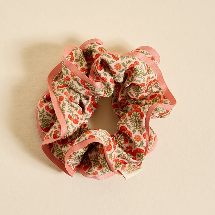 The Darling Effect's Everbloom Dusty Rose Ruffle Up Scrunchie, featuring a red and pink floral pattern with green leaves and a pink border, is shown oversized against a light beige background.