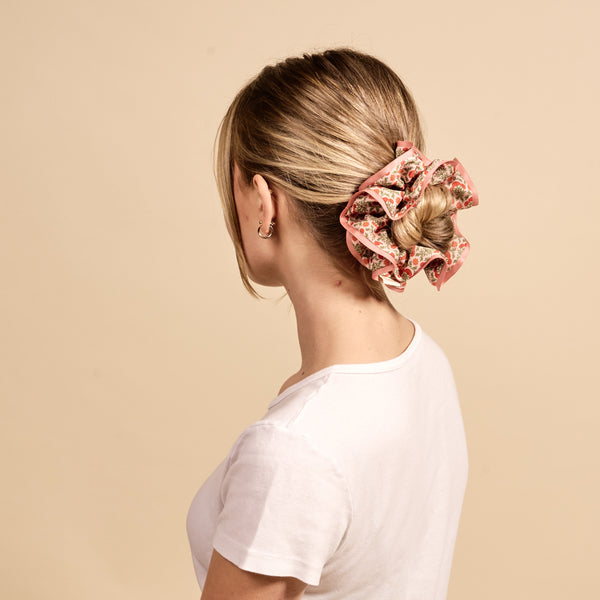 A woman with blonde hair in a low bun wears The Darling Effect's Everbloom Dusty Rose Ruffle Up Scrunchie and a white t-shirt, shown from behind against a beige background.