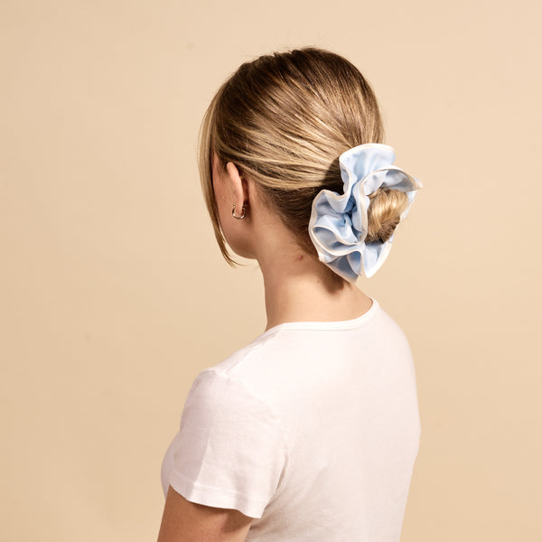 A woman with blonde hair in a low bun, styled with The Darling Effect's Light Blue Ruffle Up Scrunchie for a gentle hold, is seen from behind against a beige background. She wears a white t-shirt and small hoop earrings.