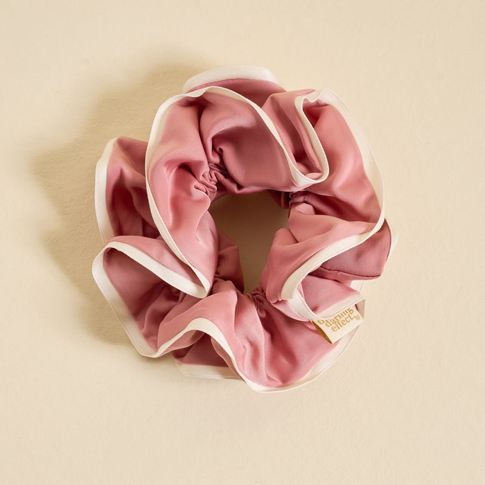 The Darling Effect’s Rose Ruffle Up Scrunchie is an oversized pink satin hair accessory with white trim, arranged in a circular shape on a light beige background. Gentle on hair, it features a small fabric tag.