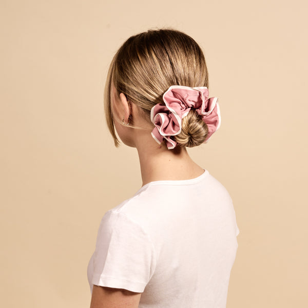 A woman with straight blonde hair styled in a low bun secured with The Darling Effect's Rose Ruffle Up Scrunchie is seen from behind, wearing a white t-shirt against a beige background.