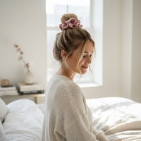 A woman with blonde hair in a bun, tied with The Darling Effect’s Rose Ruffle Up Scrunchie, sits smiling on a bed in a bright, cozy bedroom. She wears a cream sweater as sunlight streams through the window behind her.