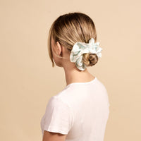 A woman with light brown hair in a low bun, secured with The Darling Effect’s Something Blue Ruffle Up Scrunchie, wears a plain white t-shirt and is shown from behind against a beige background.