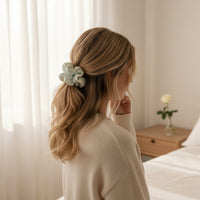 A woman with wavy blonde hair styled with The Darling Effect’s Something Blue Ruffle Up Scrunchie sits on a bed in a softly lit bedroom, wearing a cream sweater. A vase with a single flower rests on the wooden bedside table.