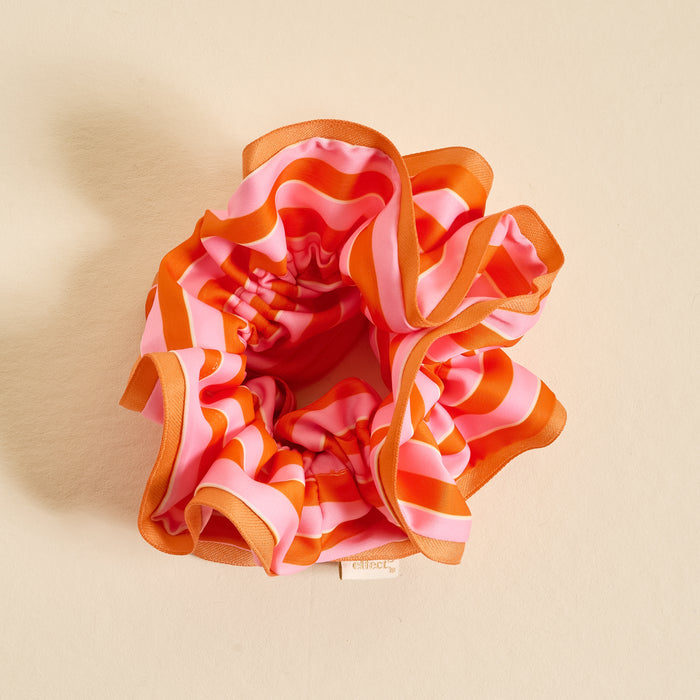 The Sicily Stripes Pink Ruffle Up Scrunchie by The Darling Effect, with pink, red, and orange wavy stripes and a ruffled design, rests on a cream surface. A fabric tag displays the brand name on this stylish accessory.