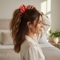 A woman with wavy brown hair, in a cream blouse, stands in a sunlit bedroom adjusting her Sicily Stripes Pink Ruffle Up Scrunchie from The Darling Effect. The room has a made bed, pillows, and a plant on the table in the background.
