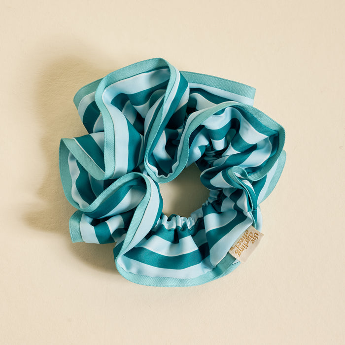 The Darling Effect’s Sicily Stripes Teal Ruffle Up Scrunchie is an oversized blue and teal striped hair accessory with a small beige tag, shown arranged in a loose circle on a light cream background.