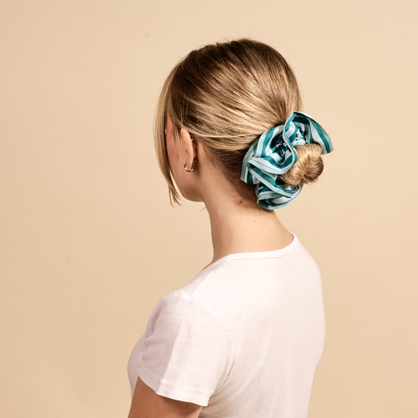 A woman with blonde hair in a low bun, secured with The Darling Effect's Sicily Stripes Teal Ruffle Up Scrunchie, wears a white t-shirt and faces away from the camera against a beige background.