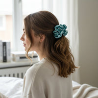 A woman with wavy brown hair sits on a bed, facing left. She wears a cream top and her hair is styled in a loose ponytail with The Darling Effect’s Sicily Stripes Teal Ruffle Up Scrunchie. Soft natural light streams through the window behind her.