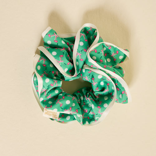 The Darling Effect’s Tee Time Ruffle Up Scrunchie, a green oversized hair accessory with white polka dots, pink bows, and white trim, sits on a light beige surface. It features ruffled details and a small attached fabric tag.