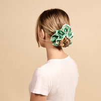 A blonde woman with her hair in a bun wears The Darling Effect's Tee Time Ruffle Up Scrunchie, a large green polka-dot ruffled accessory.