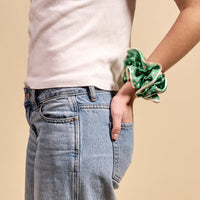 A person in a white t-shirt and blue jeans stands with one hand in their pocket, wearing The Darling Effect’s Tee Time Ruffle Up Scrunchie—a green polka dot oversized accessory—on their wrist against a beige background.