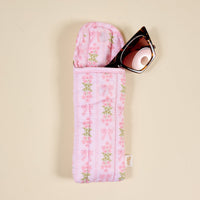 A pair of sunglasses partially tucked into The Darling Effect's Oh So Fancy Pink Puffy Sunglasses Case, featuring a floral and bow pattern with a magnetic flap closure, resting on a beige surface.