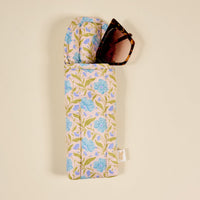 A pair of brown tortoiseshell sunglasses is partially tucked into The Darling Effect’s Poppin' Petals Aqua Puffy Sunglasses Case, featuring a blue and green floral pattern and magnetic flap closure, resting on a light background.