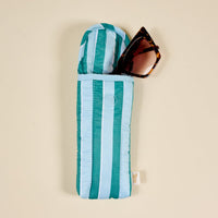 A pair of brown cat-eye sunglasses is partly tucked into The Darling Effect Sicily Stripes Teal Puffy Sunglasses Case, featuring teal and light blue stripes with a magnetic flap closure, set against a beige background.