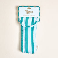 The Sicily Stripes Teal Puffy Sunglasses Case by The Darling Effect is shown in its packaging. This stylish case features teal and blue stripes, a magnetic flap closure, and a fabric tag for secure eyeglasses protection.