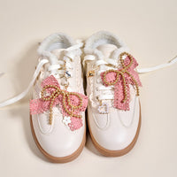 White sneakers with fluffy ankle lining, adorned with The Darling Effect's Adore Me Dazzle Shoe Charm Pack: large pink bows, gold beaded sneaker charms, and white flower accents on a light beige background.