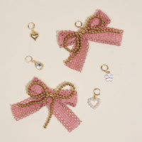 The Darling Effect's Adore Me Dazzle Shoe Charm Pack features two pink fabric bows with gold bead trim and five gold charms—a heart, teardrop gem, white flower, and two heart-shaped designs—arranged on a light surface.