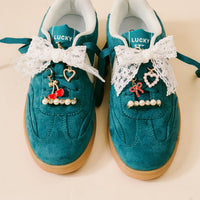 Teal suede Lucky Step sneakers adorned with white lace bows and the-darling-effect’s Cherry Twist Dazzle Shoe Charm Pack, featuring gold charms like cherries, hearts, red bows, and pearl-rhinestone bars for unique style.